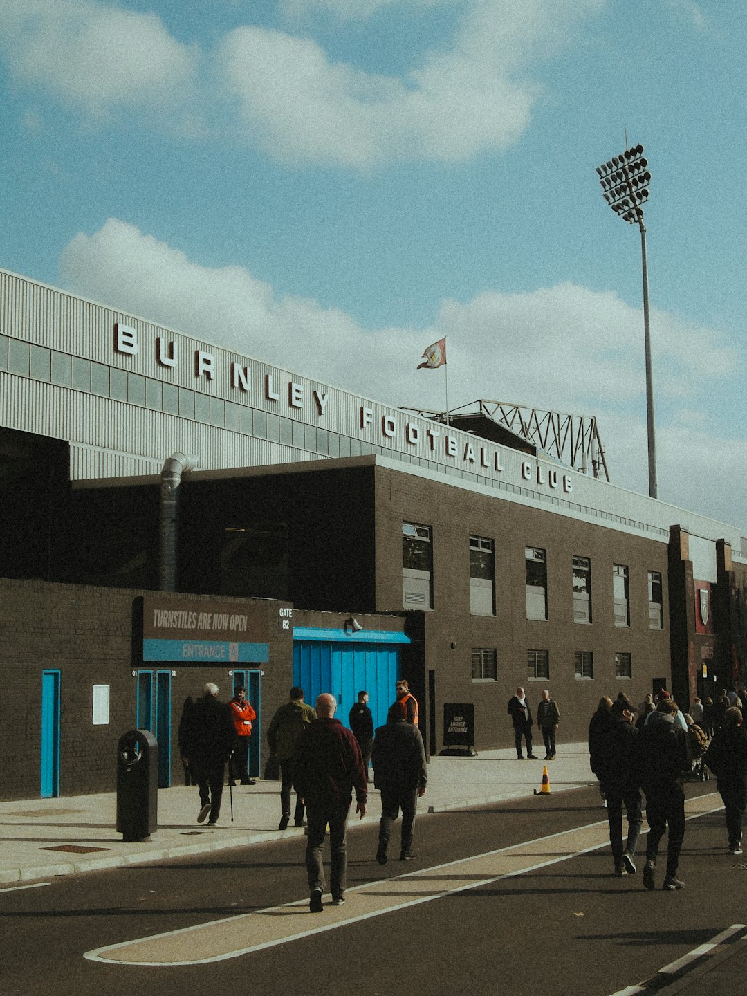 Fans walk toward blue stadium gates on a sunny day.