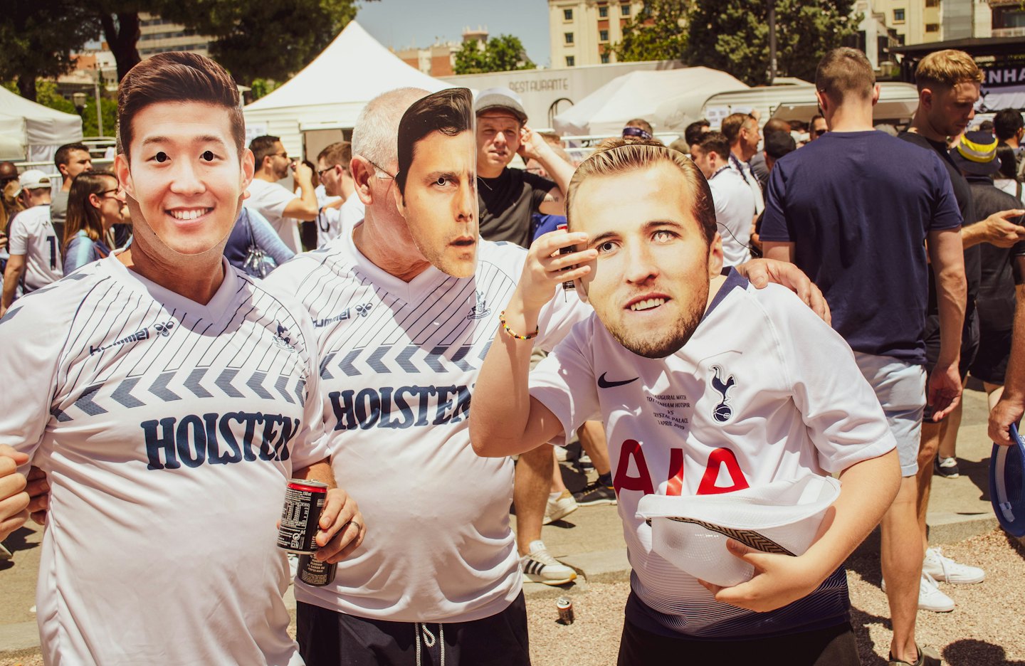 Three men hold up player face masks at a sunny outdoor fan event.