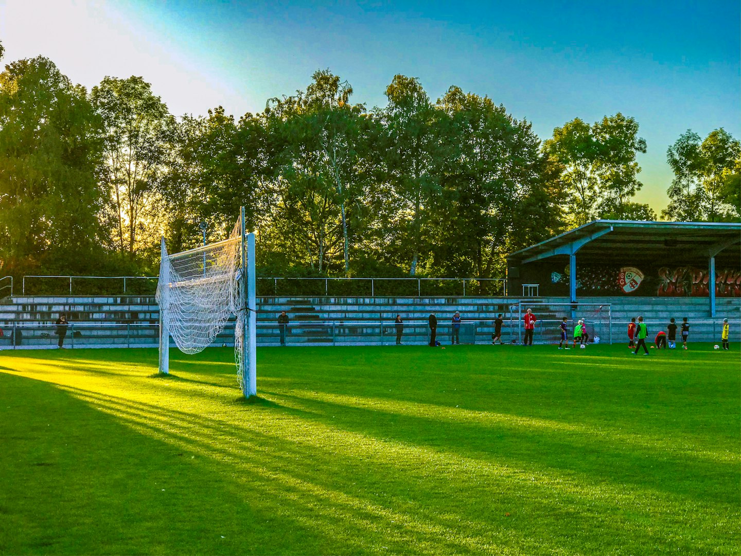 Children play soccer on a sunny field near goal net.